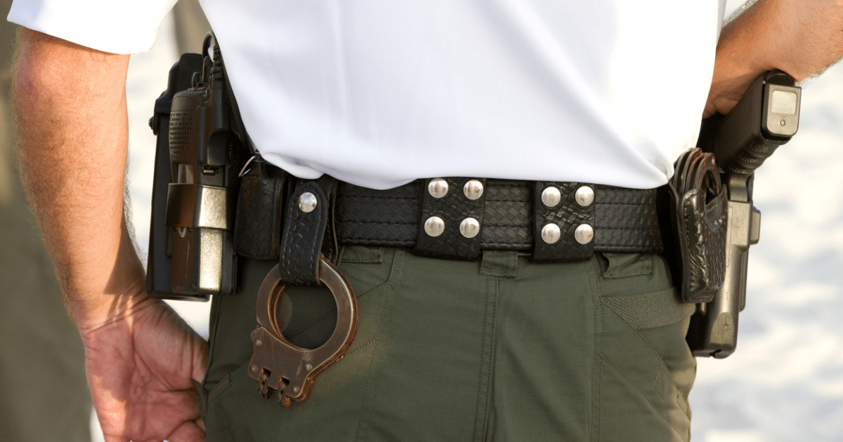 A police detective in green pants wearing a black duty belt with handcuffs and black belt keepers.