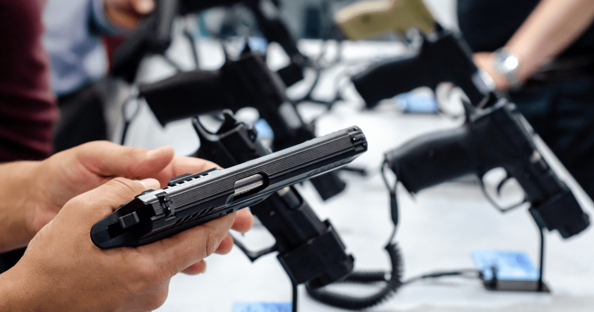 A person holding a handgun with several handguns on racks in the background.